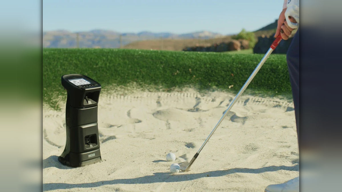 Uneekor Eye Mini launch monitor set up in a sand bunker outdoors as a golfer hits a practice shot, with mountains in the background