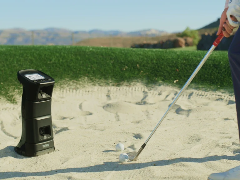 Uneekor Eye Mini launch monitor set up in a sand bunker outdoors as a golfer hits a practice shot, with mountains in the background