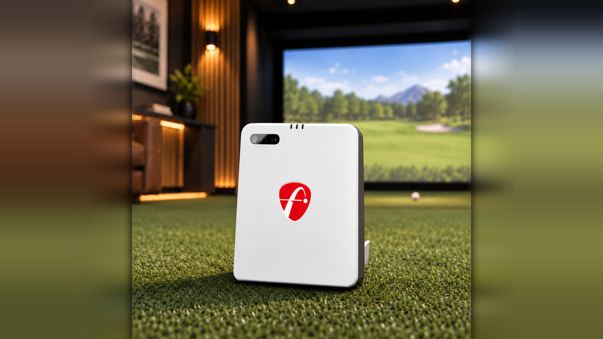 White FlightScope Mevo Gen 2 launch monitor standing on turf in a home simulator room, with a golf ball nearby and a course projected on the screen behind it