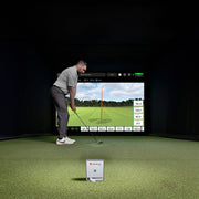 Man practicing golf swings in a simulator room with a screen displaying a golf course using his Mevo Gen2 Launch Monitor