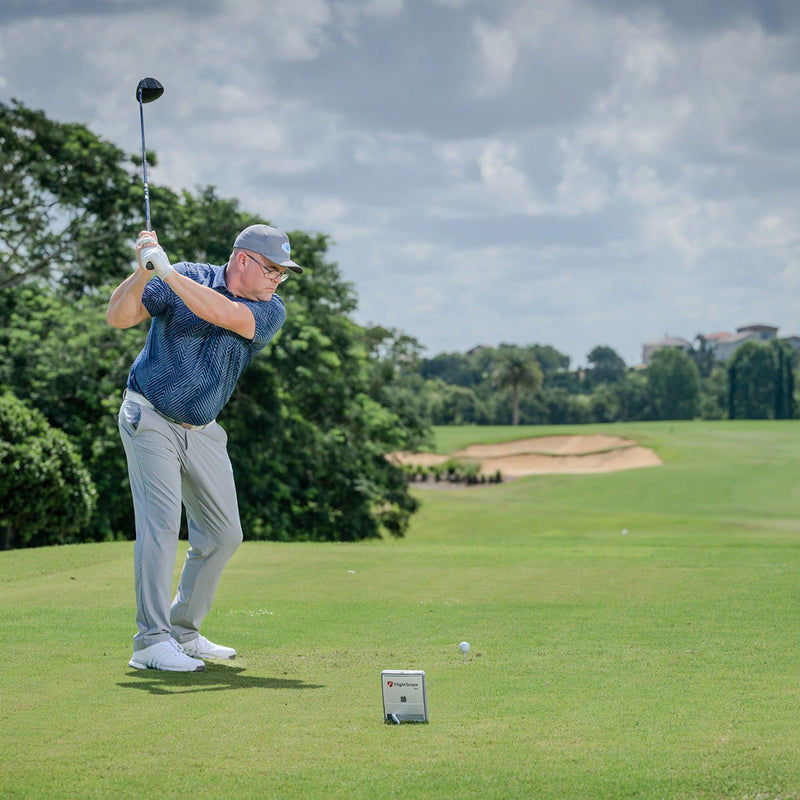 Man playing golf with a Mevo Gen2 Launch Monitor on a green course with trees and sky in the background