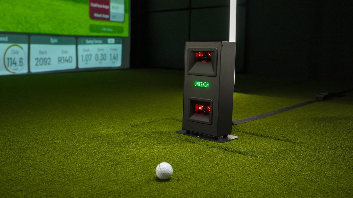 Uneekor Eye Mini Lite unit on turf with illuminated logo and sensors, golf ball nearby and shot data screen in the background