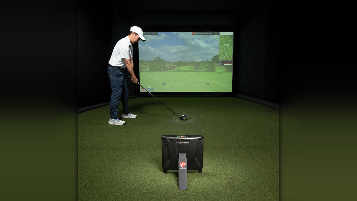 A golfer indoors in a home simulator bay addressing his ball, with the FlightScope X3C launch monitor set up behind him on turf and a golf course projected on the screen ahead