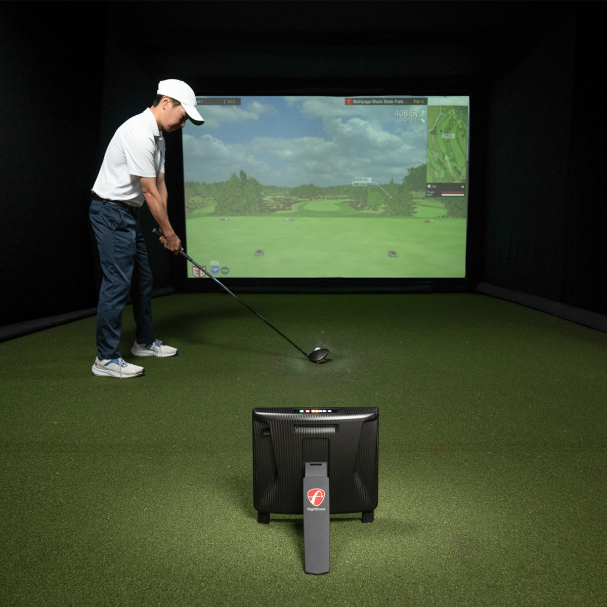 A golfer indoors in a home simulator bay addressing his ball, with the FlightScope X3C launch monitor set up behind him on turf and a golf course projected on the screen ahead