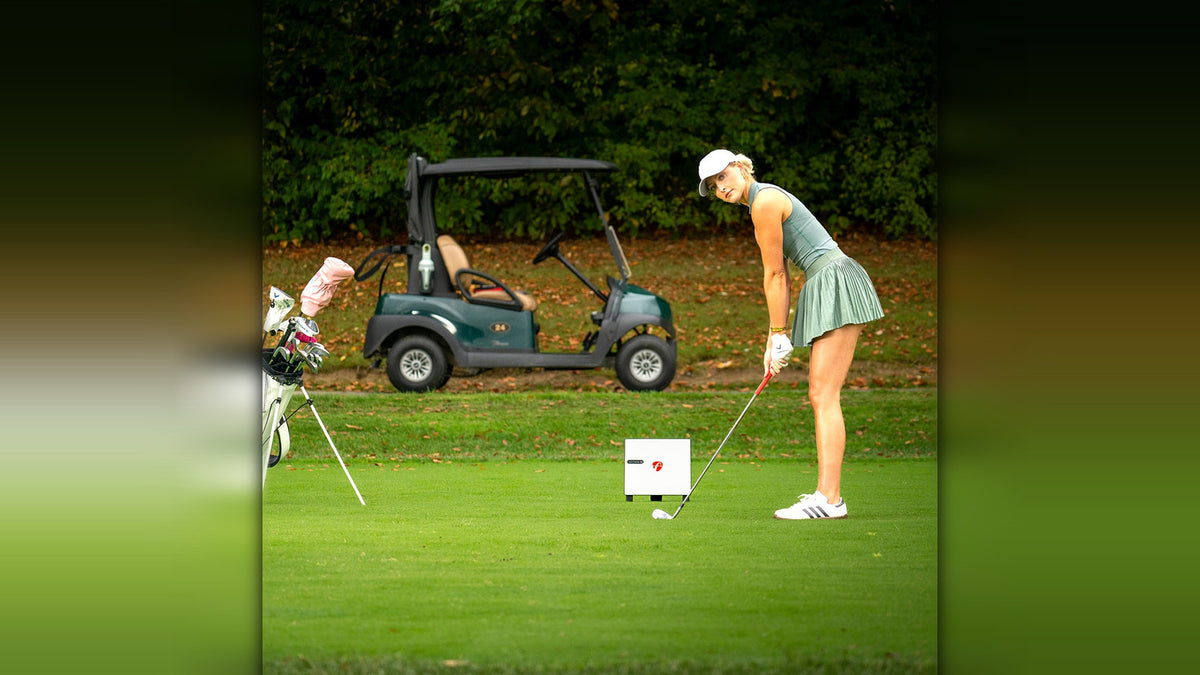 A female golfer addressing her ball on an outdoor putting green with the small white FlightScope X3C monitor placed on the grass beside her, a golf cart and bag of clubs nearby