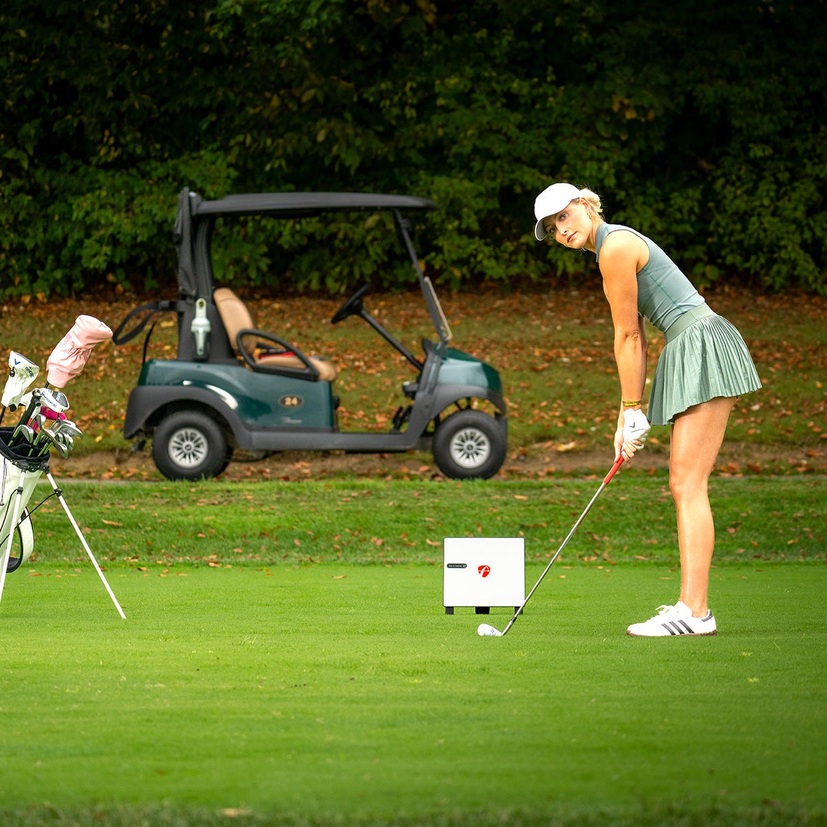 A female golfer addressing her ball on an outdoor putting green with the small white FlightScope X3C monitor placed on the grass beside her, a golf cart and bag of clubs nearby