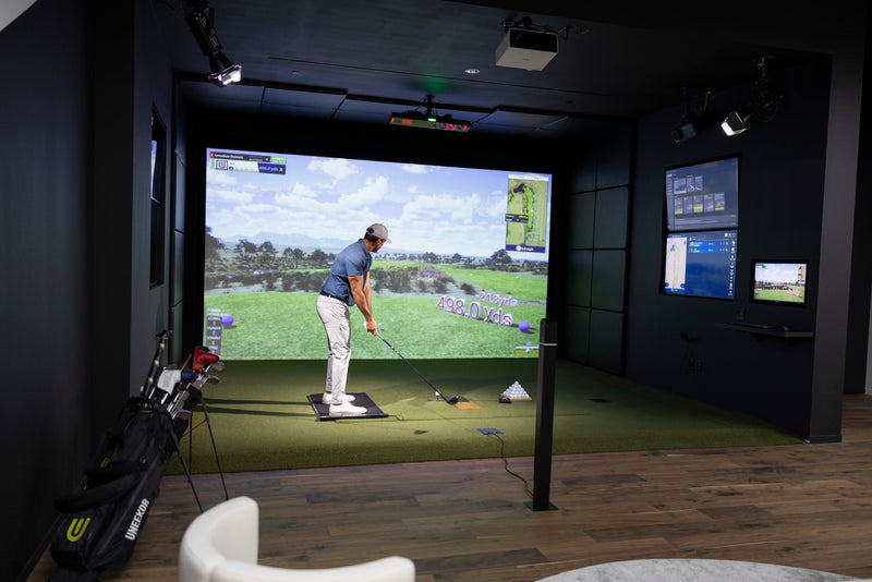 Golfer swinging in a Uneekor showroom golf simulator with the EYE XO2 mounted overhead and live data screens on the walls