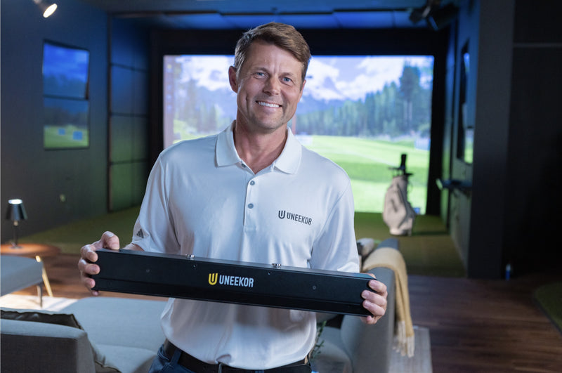 Uneekor team member holding the EYE XR launch monitor in a home golf simulator room